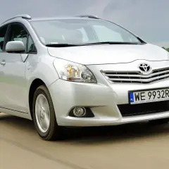 Srebrna Toyota Verso jedzie po drodze, otoczona zielonymi polami.