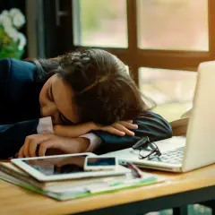 Kobieta w garniturze śpi na biurku, obok laptopa i tabletu. Może to być objaw narkolepsji.
