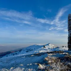 Kamienny obelisk na szczycie ośnieżonej góry, przypominający wielką rawkę. W tle błękitne niebo z chmurami i pasma górskie.