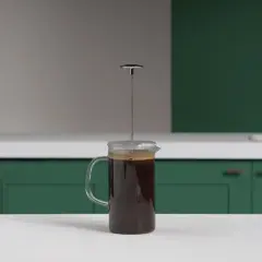 En kaffepress med mörkt kaffe redo att pressas. Köksbänk med gröna skåp och en kran i bakgrunden.