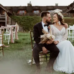 Para młoda siedzi na krzesłach na tle drewnianych budynków i białych krzeseł.