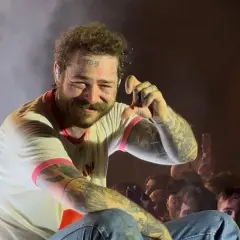 Post Malone na koncercie, uśmiecha się i pokazuje gest ręką.