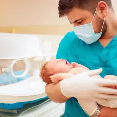 Lekarz w maseczce i rękawiczkach trzyma noworodka. To obrazek o tematyce neonatologia co to, pokazujący troskę o najmniejszych.