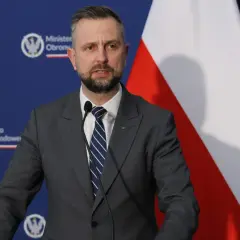 Mężczyzna w garniturze, minister obrony narodowej, przemawia przy mikrofonie na tle flagi Polski i logo ministerstwa.