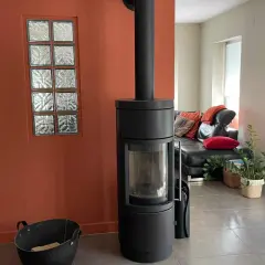 Un poêle moderne noir se dresse devant une peinture murale ocre, à côté d'une fenêtre en verre dépoli. Un canapé en cuir noir et des coussins colorés sont visibles au loin.