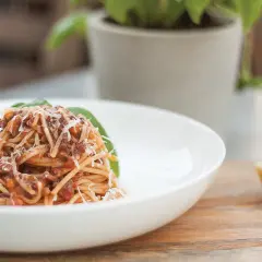 Pyszne spaghetti bolognese przepis oryginalny, podane na białym talerzu, posypane parmezanem. Obok kawałek sera i bazylia.
