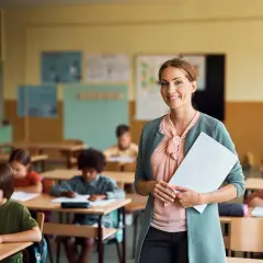 Lächelnde Lehrerin mit Unterlagen in einem Klassenzimmer voller konzentrierter Kinder. Sie fragt sich: "Soll ich Lehrer werden?