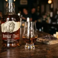 Butelka burbona Buffalo Trace, kieliszek z bursztynowym płynem, kawałki czekolady i sera na drewnianej desce.