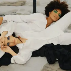 Two women in white shirts and pants, one with green eyeshadow, the other with blue. The fabric looks smooth, suggesting lyocell does not wrinkle easily.