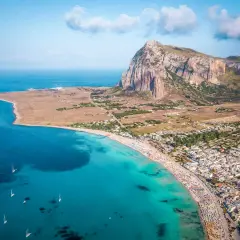 Aerial view of a crowded beach and town, showcasing one of the best beach towns in Sicily with turquoise waters and a dramatic mountain backdrop.