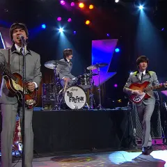 Una cover band sul palco, con abiti grigi e chitarre, ricorda i Fab Four.