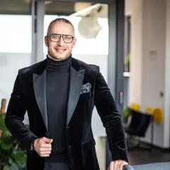 Mężczyzna w czarnym garniturze i okularach stoi w nowoczesnym biurze.
