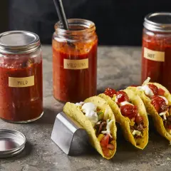 Tre burkar med tacosås, märkta "Mild", "Medium" och "Hot", står bredvid färdiga tacos. Perfekt för att hitta ditt tacosås recept.