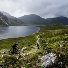 Two cyclists navigate a rocky trail with a vast lake and dramatic mountains behind them, showcasing the rugged beauty of Scotland bike tours.