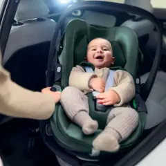 Uśmiechnięte dziecko w foteliku samochodowym. Ręka rodzica pokazuje, jak wypiąć fotelik z bazy isofix.