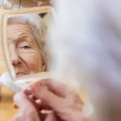 Eine ältere Frau betrachtet ihr Gesicht im Spiegel. Eine Creme steht daneben. Sie sucht nach Wegen aus der Einsamkeit, indem sie sich selbst pflegt.