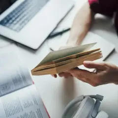 Osoba przegląda książkę, przygotowując się do napisania bibliografii. Obok leży laptop i słuchawki.