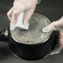 Ręce w rękawiczkach czyszczą przypalony garnek magiczną gąbką.