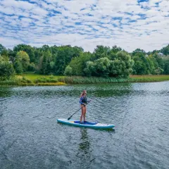 Kobieta na desce SUP na spokojnym jeziorze, w tle zielone drzewa i błękitne niebo.