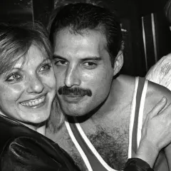 Freddie Mercury z uśmiechem, odsłaniającym jego charakterystyczne zęby, przytula się do kobiety.