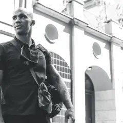 Nico Musoke, en man med tatueringar, bär en väska framför en byggnad i svartvitt.