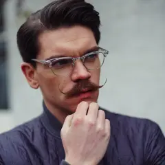Un homme au visage losange, lunettes fines et moustache élégante, réfléchit.
