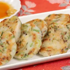 Golden-brown daikon mochi patties, studded with green onions, are served on a white platter with a small dish of dipping sauce.