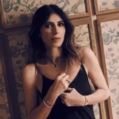 Woman in black slip dress leans against a wall with floral wallpaper.