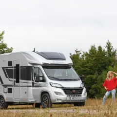 Une femme pose devant un camping-car Adria Matrix 670 SC argenté, prêt pour l'aventure.