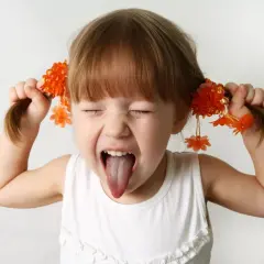 Bambina iperattiva fa una smorfia con la lingua fuori, tirando le trecce con fiori arancioni. Cosa fare?