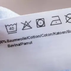 Pflegeetikett mit Waschsymbolen und dem Hinweis "100% Baumwolle". Die Symbole zeigen, dass das Kleidungsstück bei 40°C waschbar ist, nicht bleichen, bei niedriger Temperatur im Trockner trocknen und bei mittlerer Hitze bügeln.