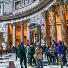 Wnętrze Panteonu w Rzymie, gdzie turyści zwiedzają, a przewodnik z czerwoną chorągiewką pokazuje detale. Bilety do Panteonu są niezbędne, by podziwiać to arcydzieło.