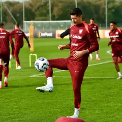 Piłkarze w bordowych strojach ćwiczą na boisku. Robert Lewandowski bawi się piłką, pokazując swoje umiejętności w najpopularniejszym sporcie na świecie.