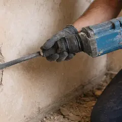 Ręka w rękawicy używa młota udarowego, by skuć tynk do cegły.