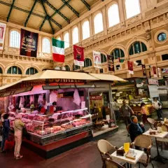 Interno di un vivace mercato delle vettovaglie, con banchi di carne, prodotti freschi e persone che fanno acquisti.