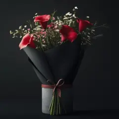 Un bouquet de callas rouges et de petites fleurs blanches, emballé dans du papier noir et noué avec un ruban de cuir. Une fleur pour homme élégante et moderne.
