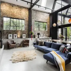 A spacious living area with exposed brick walls, concrete floors, and industrial interior design elements. A blue sectional sofa, leather armchairs, and a large dining table create a cozy yet modern atmosphere.