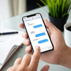 Dłoń trzyma smartfon z otwartym komunikatorem. Na ekranie widać rozmowę: "At work. See you today?". Obok filiżanka kawy.