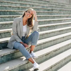 Uśmiechnięta kobieta w jasnych jeansach i marynarce siedzi na schodach. Stylowe, codzienne jasne jeansy stylizacje damskie.