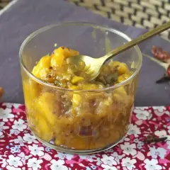 Un verre rempli de chutney de mangue, avec une cuillère dorée. Des épices comme la cannelle, le piment et le clou de girofle entourent le plat.