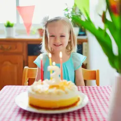 Ein strahlendes 3 Jahre altes Kind sitzt vor seinem Geburtstagskuchen mit brennenden Kerzen. Girlanden schmücken den Raum für die Feier.