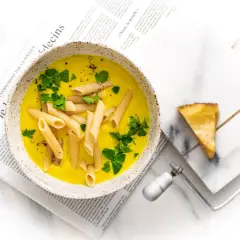 Zupa neapolitańska z makaronem penne i natką pietruszki, podana na gazecie. Obok kawałek sera i serownik.