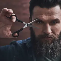 Mężczyzna z bujną brodą i nożyczkami przy oku, gotowy do modelowania brody.