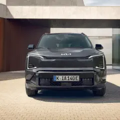 Nowoczesny, ciemnoszary Kia EV5 z charakterystycznymi światłami LED stoi przed minimalistycznym budynkiem z elementami rdzy i betonu.