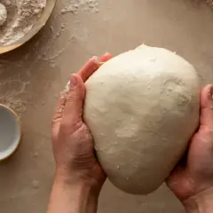 Mani che impastano un panetto soffice, ingrediente chiave per imparare come fare il lievito madre. Farina e ciotolina a lato.