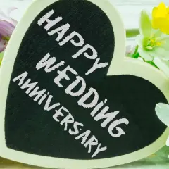 Cuore nero con scritta "Happy Wedding Anniversary" circondato da fiori bianchi, gialli e viola. Un'idea romantica per sapere dove festeggiare anniversario matrimonio.