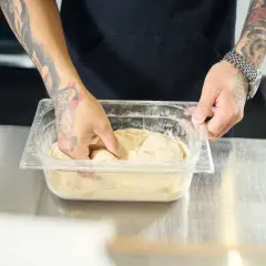 Ręce z tatuażami mieszają ciasto na pizzę w pojemniku. Dowiedz się, jak szybko rozmrozić ciasto na pizzę.