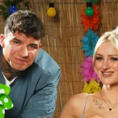 Ein Mann mit Tätowierungen und eine Frau mit blonden Haaren lächeln vor einer Bambuswand mit bunten Lichtern und Blumen. Sie sind bereit für das Sommerhaus der Stars 2025.