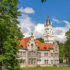 Pałac w Nakle Śląskim otoczony zielenią, z charakterystyczną wieżą i czerwonym dachem.