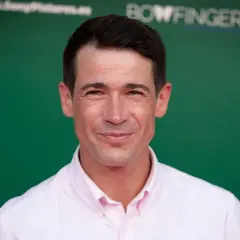 Juan José Ballesta, cuya edad se refleja en su mirada, sonríe con camisa rosa ante un fondo verde con logos.